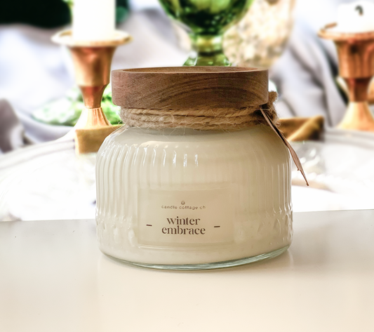 Vintage-style scented candle in glass jar with wooden lid, labeled "Winter Embrace," surrounded by elegant decor.